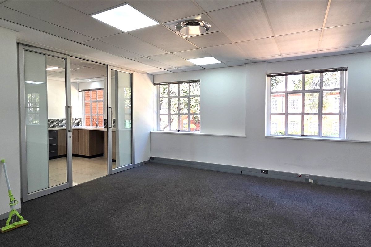 A bright, open-plan office space featuring carpeted flooring, a suspended ceiling with recessed lighting, and a sliding glass partition leading to a kitchenette area.