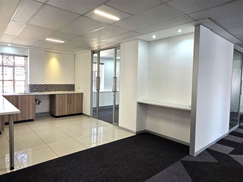 A modern, open-plan office space featuring a kitchenette area, glass partition doors, suspended ceilings, and a mix of tiled and carpeted flooring.