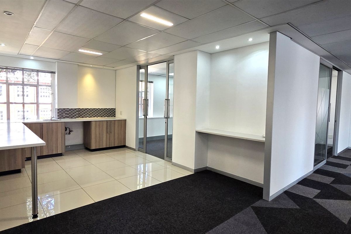 A modern, open-plan office space featuring a kitchenette area, glass partition doors, suspended ceilings, and a mix of tiled and carpeted flooring.