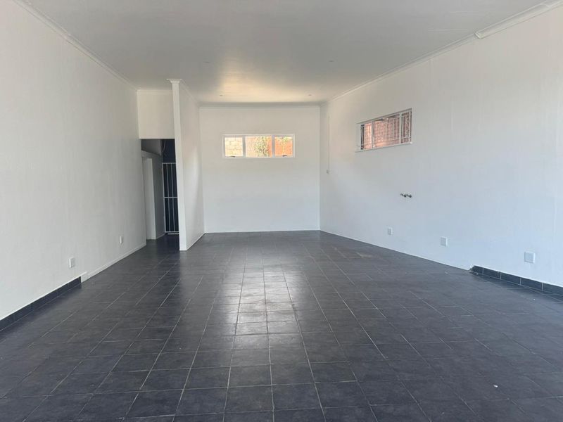 A spacious, open-plan retail area featuring neutral white walls, dark tiled flooring, and security bars on the windows.