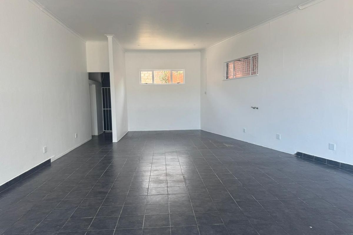 A spacious, open-plan retail area featuring neutral white walls, dark tiled flooring, and security bars on the windows.