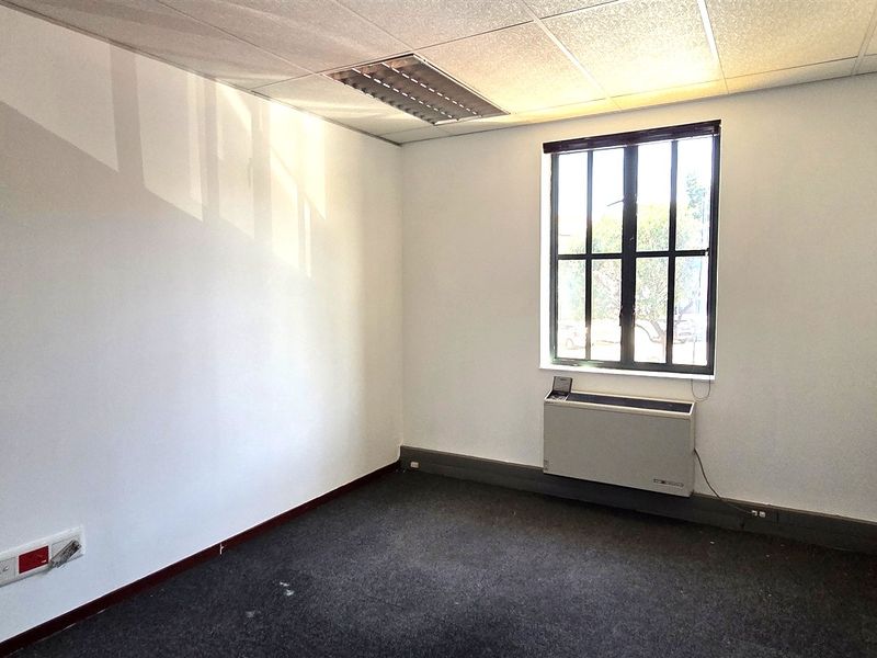 A vacant office space featuring a suspended ceiling, recessed lighting, wall-mounted HVAC unit, and carpeted flooring, providing a functional workspace environment.