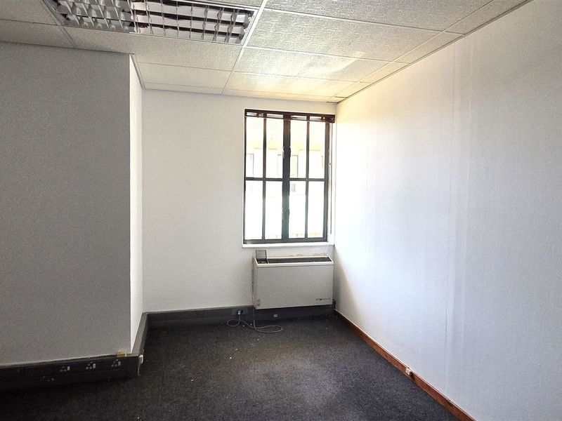 A vacant office space featuring a suspended ceiling, carpeted flooring, a wall-mounted air conditioning unit, and a window with security bars.