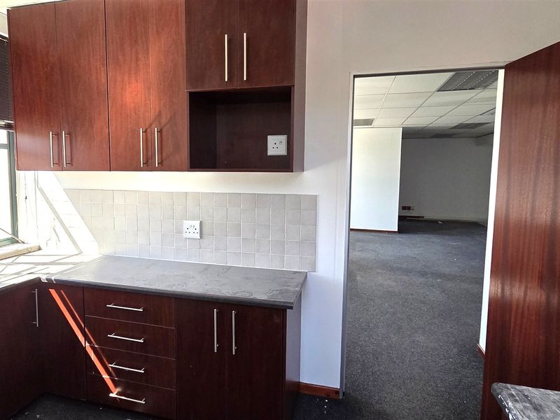 A functional office kitchenette featuring dark wood-look cabinetry, a stainless steel sink, and a tiled backsplash, leading into an open-plan office area.