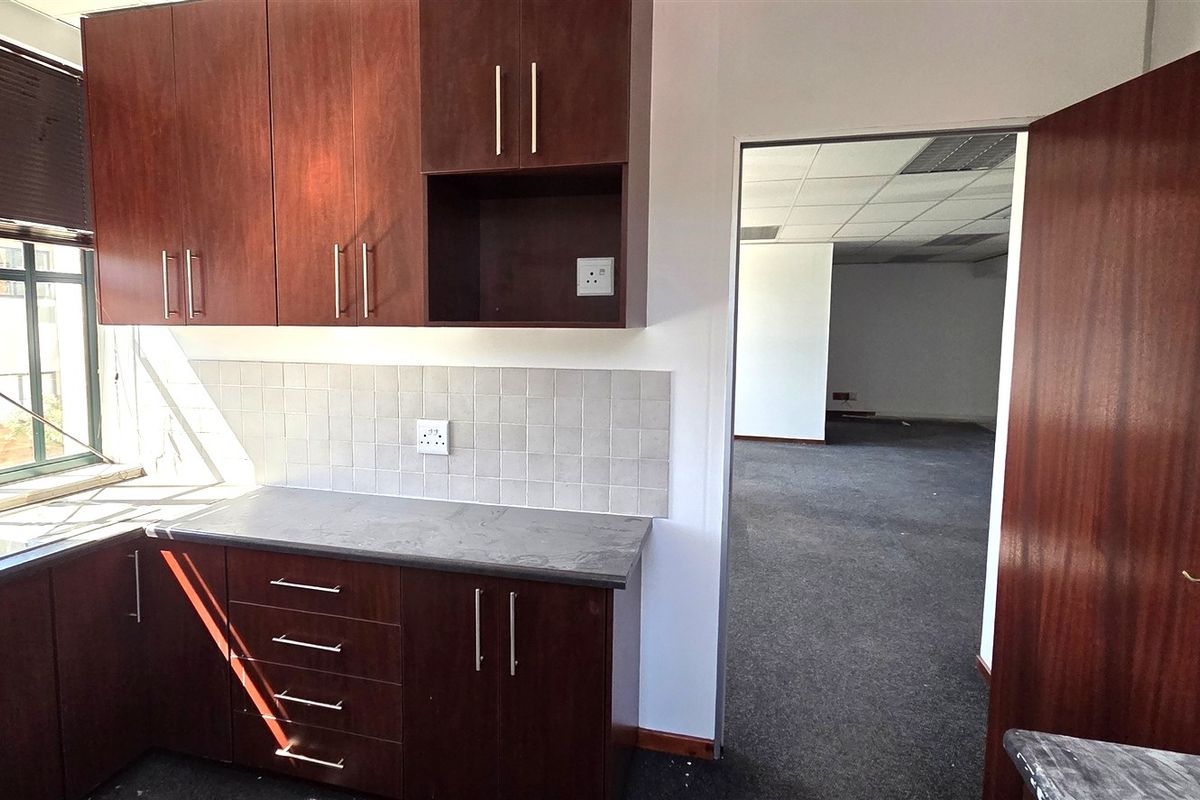 A functional office kitchenette featuring dark wood-look cabinetry, a stainless steel sink, and a tiled backsplash, leading into an open-plan office area.