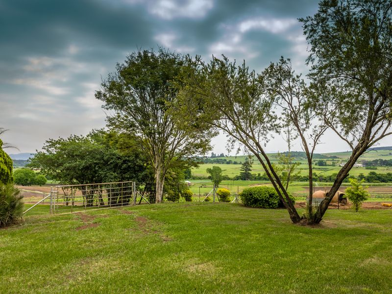 A well-maintained grassy garden area featuring mature trees, wire fencing, and a metal farm gate, overlooking expansive rural farmland.