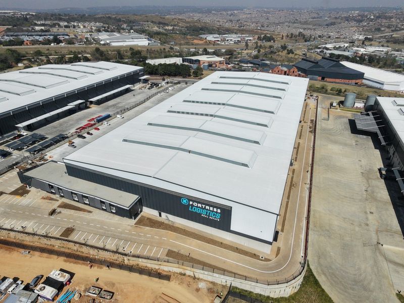 Aerial view of a large industrial warehouse facility featuring a metal roof with skylights, paved driveway, and secure perimeter fencing.