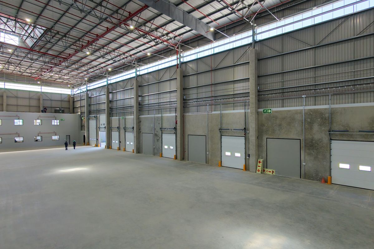 Spacious industrial warehouse featuring high ceilings, exposed steel trusses, multiple roller shutter doors, and polished concrete flooring, suitable for logistics or manufacturing operations.