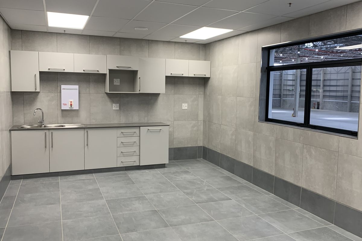 A clean, modern industrial kitchenette featuring neutral-toned cabinetry, tiled surfaces, and a window overlooking the warehouse floor.