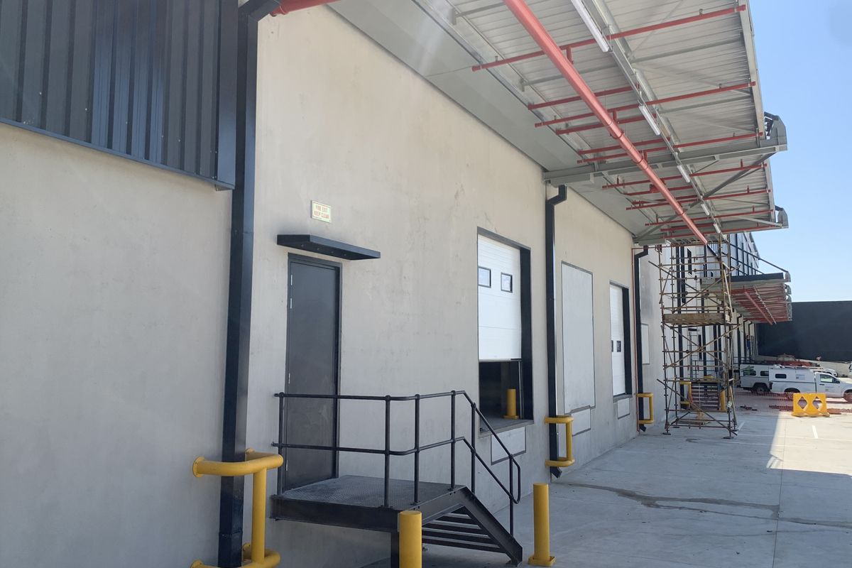 Industrial exterior view featuring a loading dock, roller shutter door, safety bollards, and overhead fire sprinkler infrastructure.