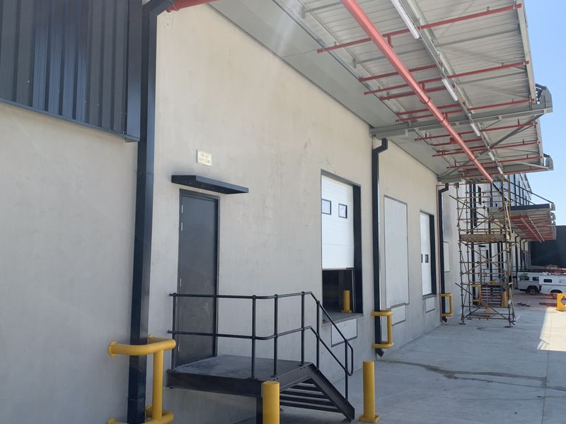 Industrial exterior view featuring a loading dock, roller shutter door, safety bollards, and overhead fire sprinkler infrastructure.