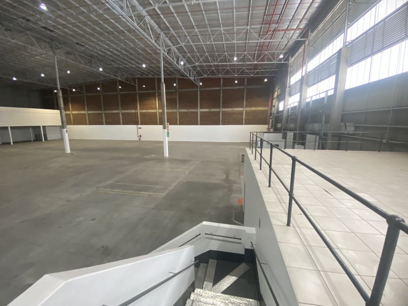 Spacious industrial warehouse featuring high ceilings with exposed steel trusses, concrete flooring, and a tiled mezzanine level accessible via a staircase.