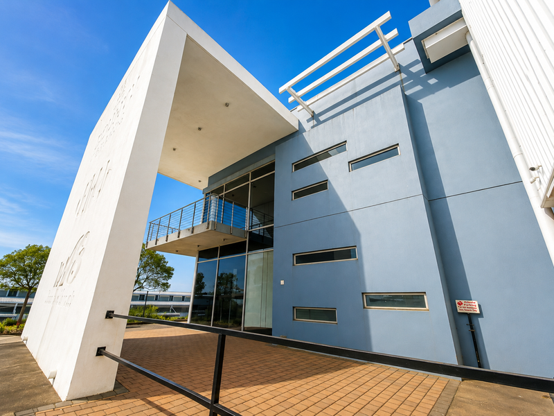 Modern industrial building facade featuring a prominent covered entrance, blue exterior walls, horizontal windows, and an elevated balcony with metal railings.