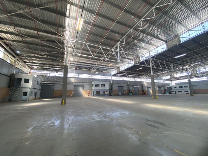 Expansive industrial warehouse space featuring high ceilings with exposed steel trusses, polished concrete flooring, multiple roller shutter doors, and integrated mezzanine office units.