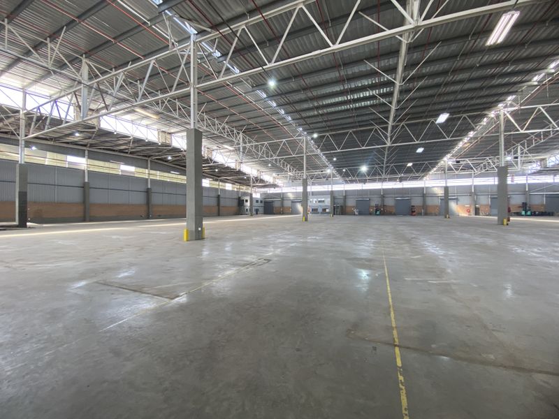 A large, open-plan industrial warehouse space featuring high-clearance steel truss roofing, polished concrete floors, and multiple roller shutter access points.