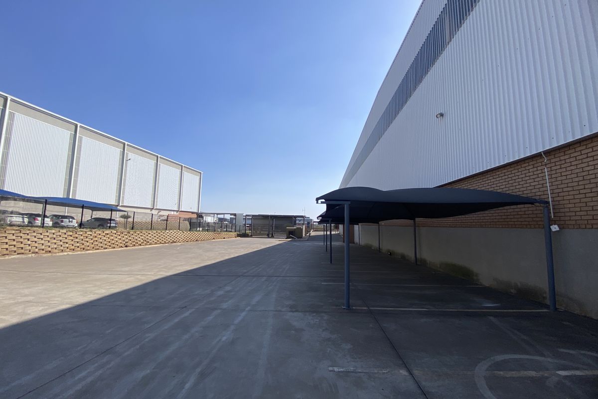 Spacious industrial yard area featuring concrete paving, covered parking bays with shade cloth structures, and large warehouse buildings with metal and brick facades.