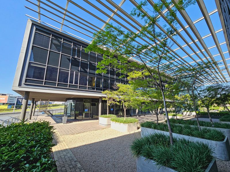 Modern office building exterior featuring a large metal pergola, extensive glass curtain walling, and well-maintained concrete planters with integrated landscaping.