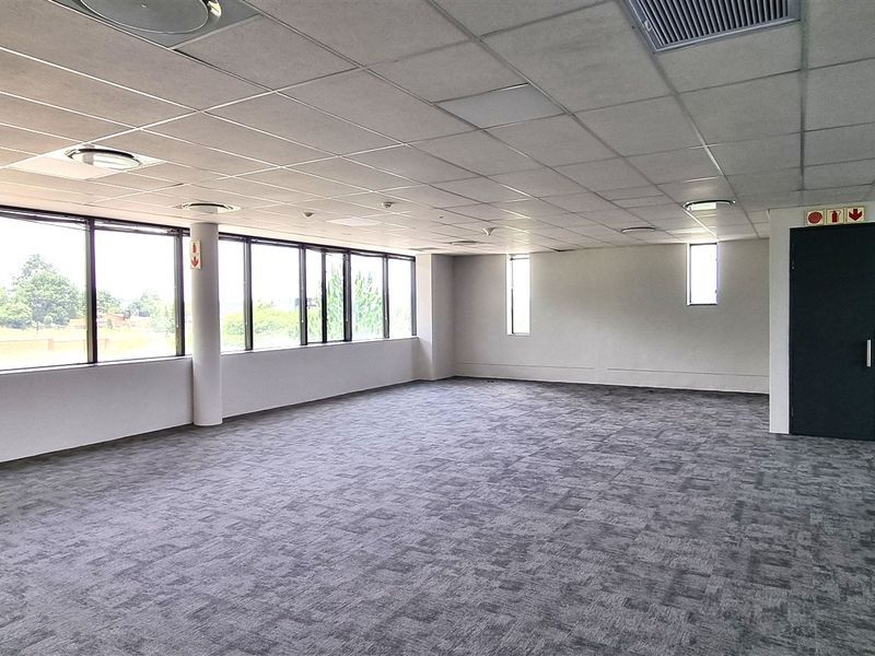 A spacious, empty open-plan office area featuring large windows, carpeted flooring, and a suspended ceiling with integrated lighting and air conditioning.