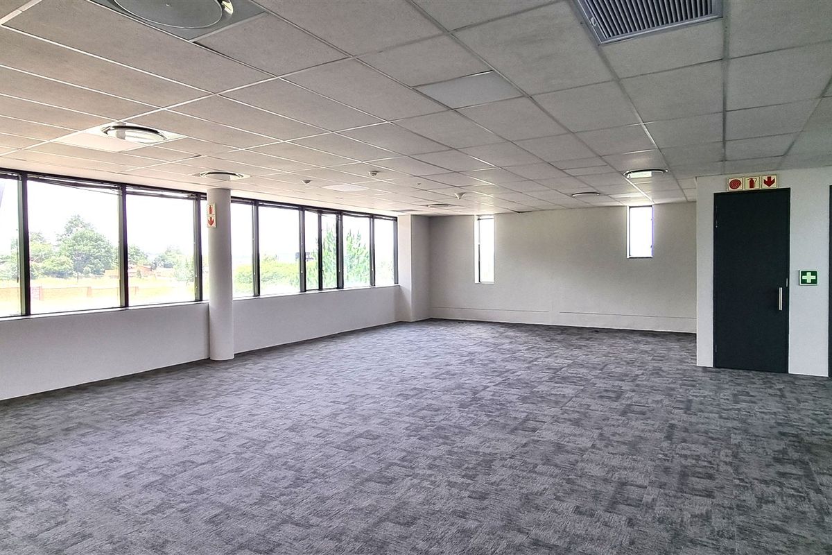 A spacious, empty open-plan office area featuring large windows, carpeted flooring, and a suspended ceiling with integrated lighting and air conditioning.