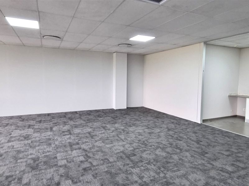 A spacious, open-plan office area featuring carpet tiles, a suspended ceiling with recessed lighting, and a small built-in kitchenette nook.