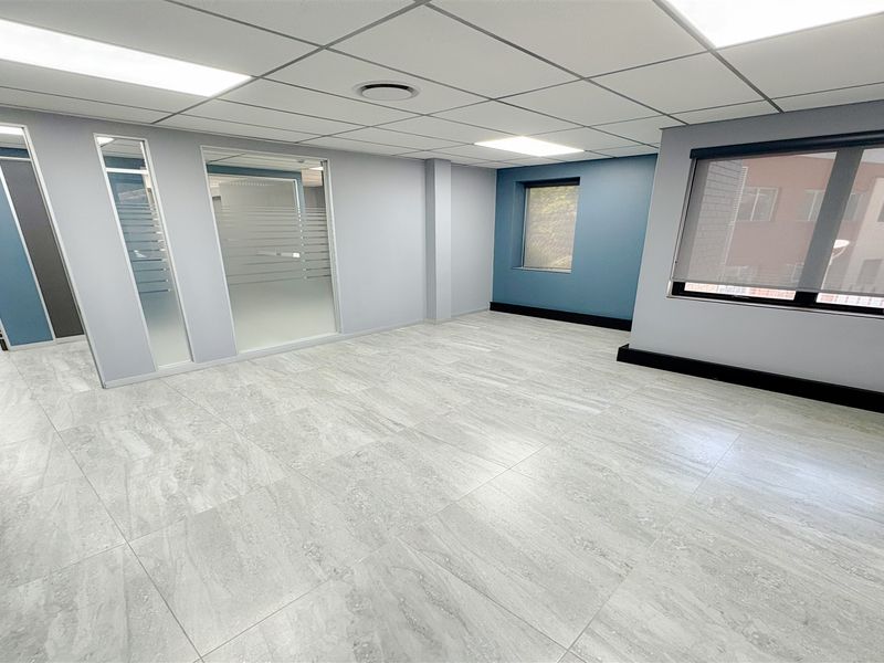 A bright, modern office space featuring neutral tiled flooring, glass partitions, and a suspended ceiling with recessed lighting.