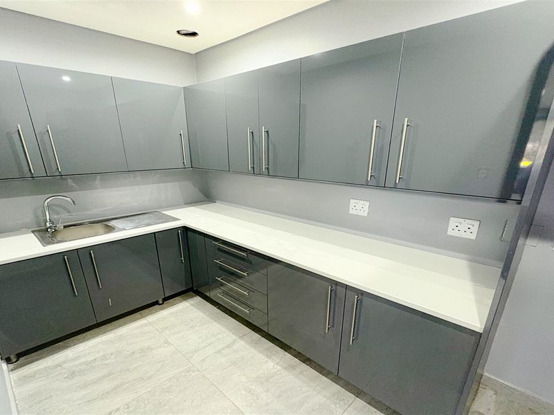 A modern, empty kitchen area featuring sleek grey built-in cabinetry, white stone countertops, a stainless steel sink, and contemporary tiled flooring.
