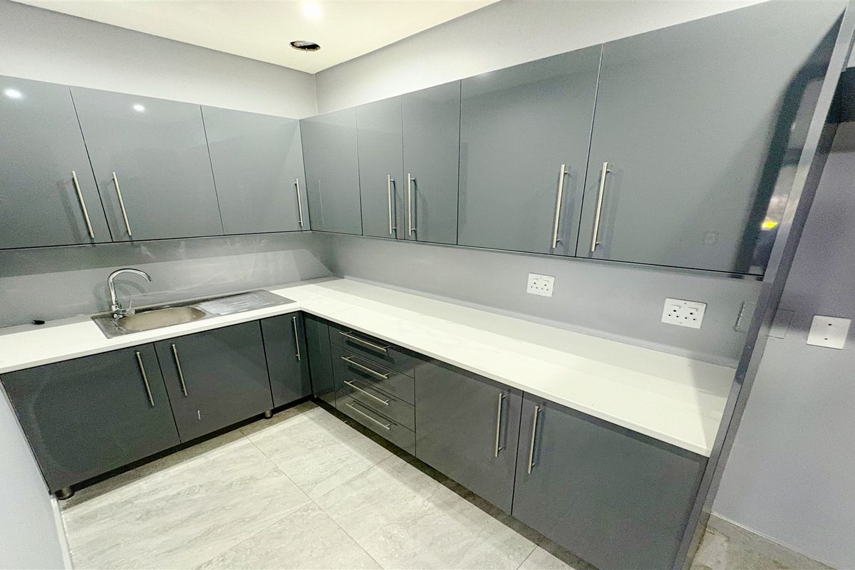 A modern, empty kitchen area featuring sleek grey built-in cabinetry, white stone countertops, a stainless steel sink, and contemporary tiled flooring.