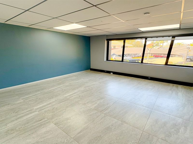 A bright, empty office space featuring modern tiled flooring, a suspended ceiling with recessed lighting, and large windows providing natural light.