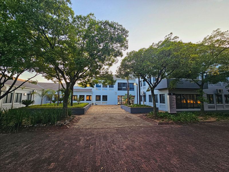 A well-maintained office complex exterior featuring a paved entrance courtyard, mature trees, and landscaped garden beds.