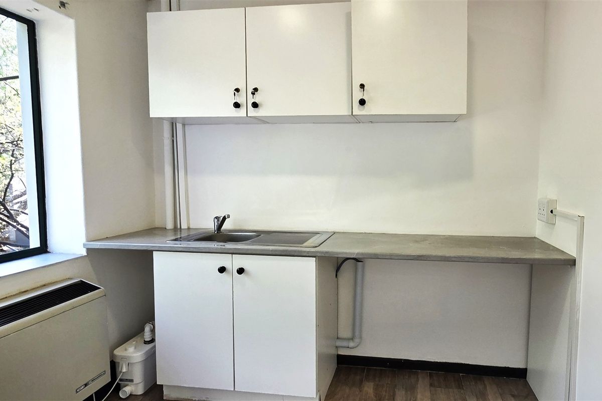 A functional office kitchenette featuring white built-in cabinetry, a stainless steel sink, laminate countertop, and a large window providing natural light.