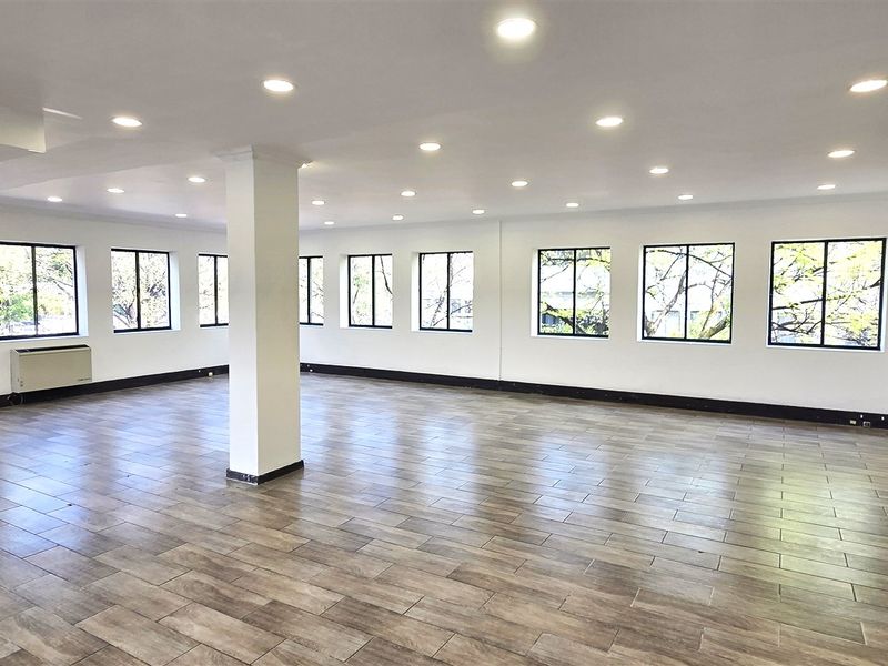 A spacious, open-plan office area featuring wood-look tile flooring, recessed lighting, and multiple windows providing ample natural light.