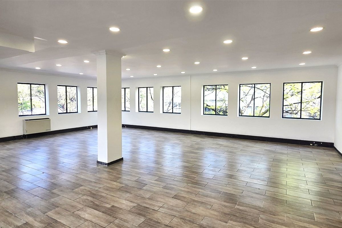 A spacious, open-plan office area featuring wood-look tile flooring, recessed lighting, and multiple windows providing ample natural light.