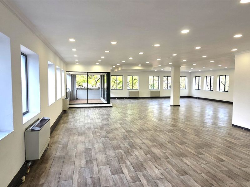 Spacious, open-plan office area featuring modern wood-look tile flooring, recessed lighting, and large windows providing ample natural light and balcony access.