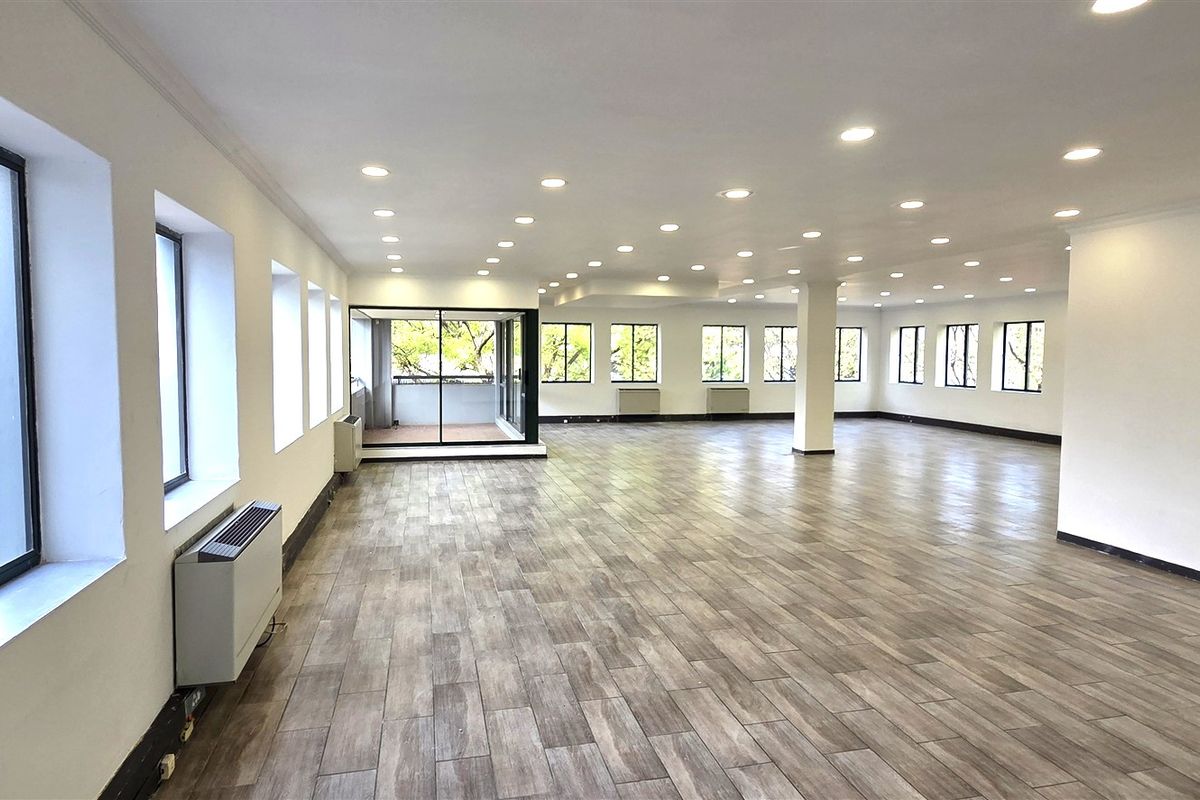 Spacious, open-plan office area featuring modern wood-look tile flooring, recessed lighting, and large windows providing ample natural light and balcony access.