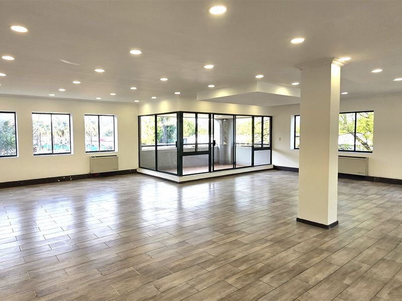 Spacious, open-plan office area featuring wood-look tile flooring, recessed lighting, HVAC units, and a central glass-enclosed meeting room or private office space.