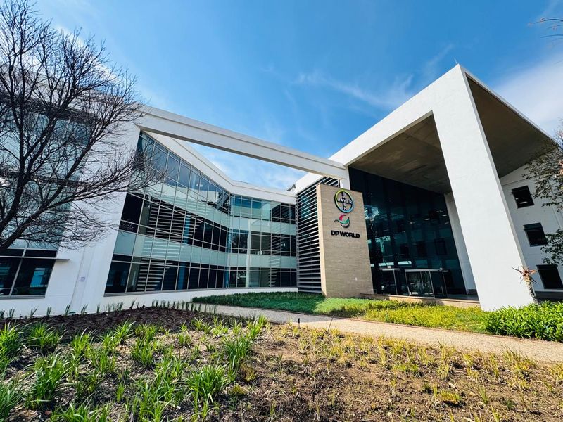 Modern commercial office building exterior featuring a prominent glass facade, concrete structural elements, and well-maintained landscaped garden beds at the entrance.