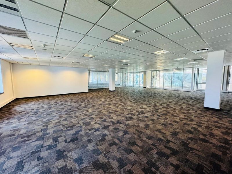 A spacious, open-plan office area featuring modern carpet tiles, a suspended ceiling with recessed lighting, and extensive glass partitioning for natural light.