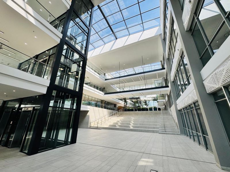 Modern office building atrium featuring a glass elevator, expansive skylight, and tiered concrete staircase, creating a bright and professional multi-level entrance space.