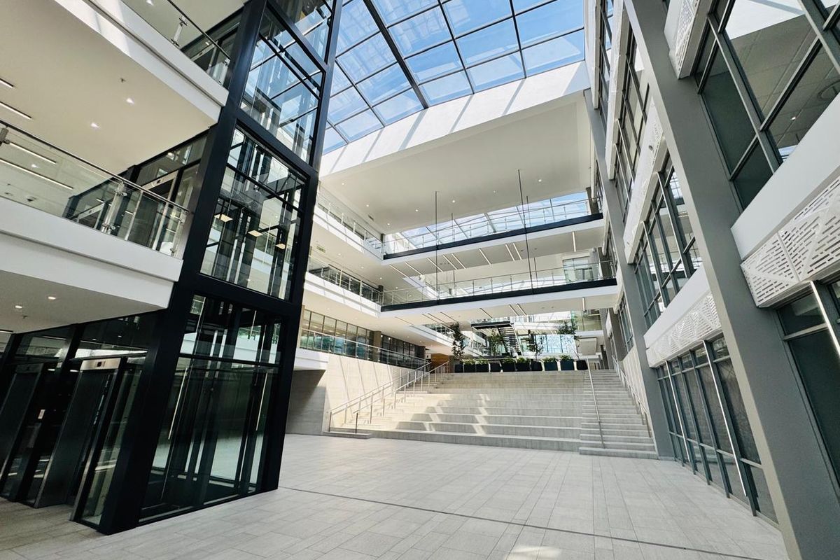 Modern office building atrium featuring a glass elevator, expansive skylight, and tiered concrete staircase, creating a bright and professional multi-level entrance space.