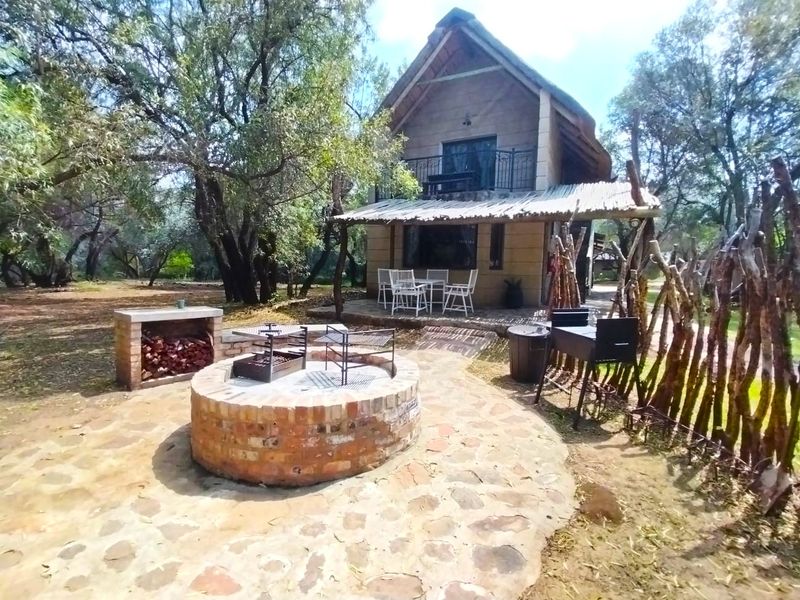 A rustic farm-style dwelling featuring a thatched roof, stone-paved patio, built-in brick fire pit, and a balcony overlooking a wooded garden area.