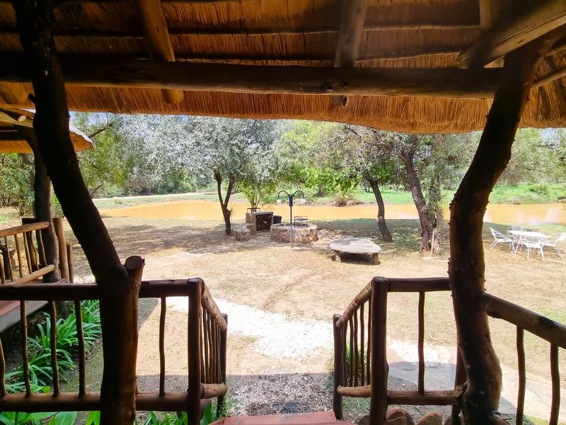 A rustic covered patio with a thatched roof and timber railings, overlooking a natural garden area with a built-in braai and fire pit.