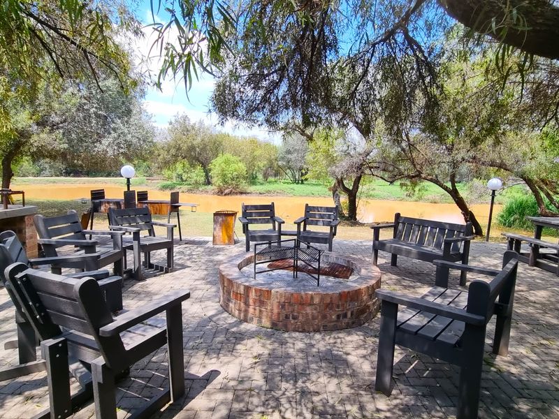 Outdoor entertainment area featuring a brick fire pit and built-in braai, set on a paved patio overlooking the riverbank.