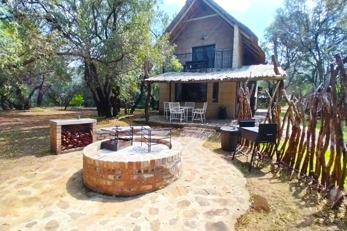 A rustic farm-style dwelling featuring a thatched roof, stone-paved patio, built-in brick fire pit, and a balcony overlooking a wooded garden area.