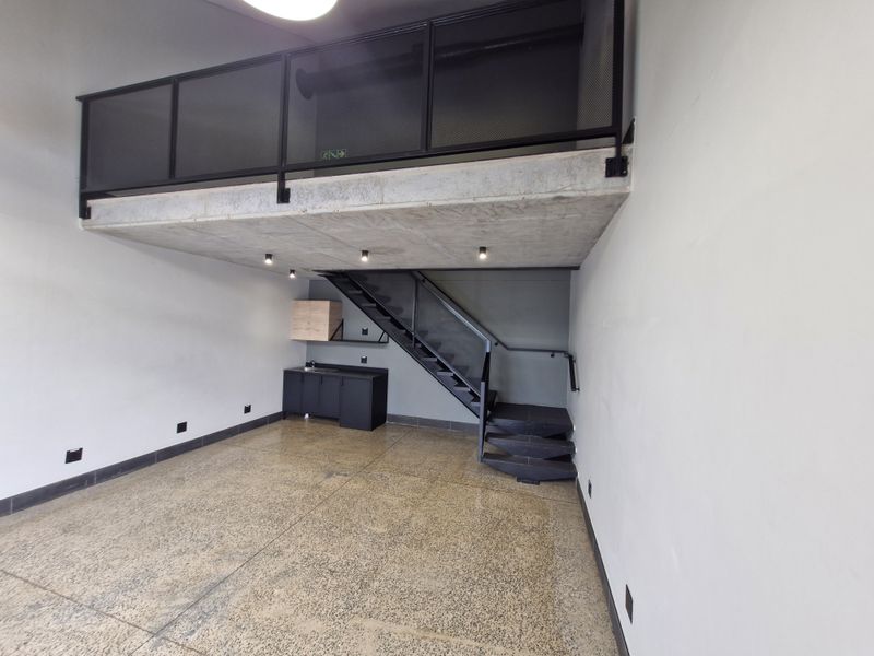Modern open-plan retail or office space featuring a mezzanine level, polished concrete floors, industrial metal staircase, and a compact kitchenette area.