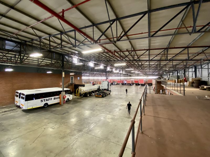 Large industrial warehouse space featuring high exposed steel roof trusses, fire sprinkler systems, concrete flooring, and an elevated mezzanine viewing area.