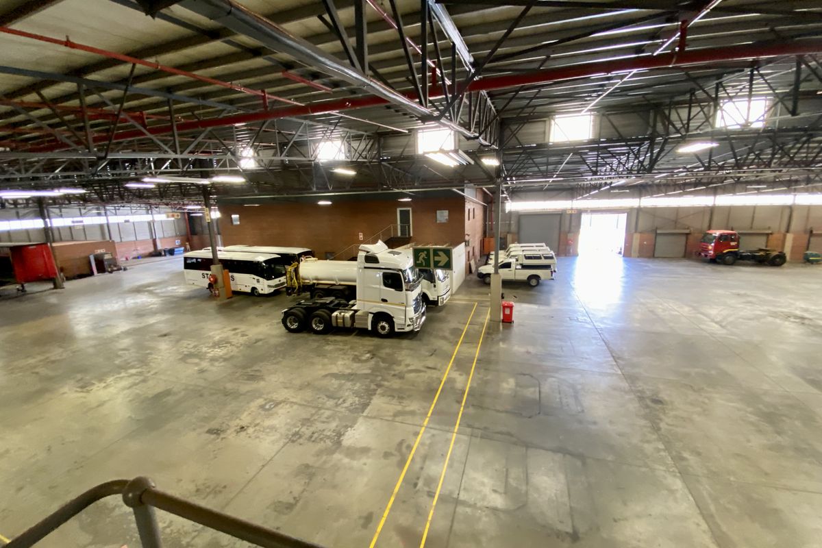 Spacious industrial warehouse featuring high ceilings with exposed steel trusses, concrete flooring, fire sprinkler systems, and large roller shutter access doors.