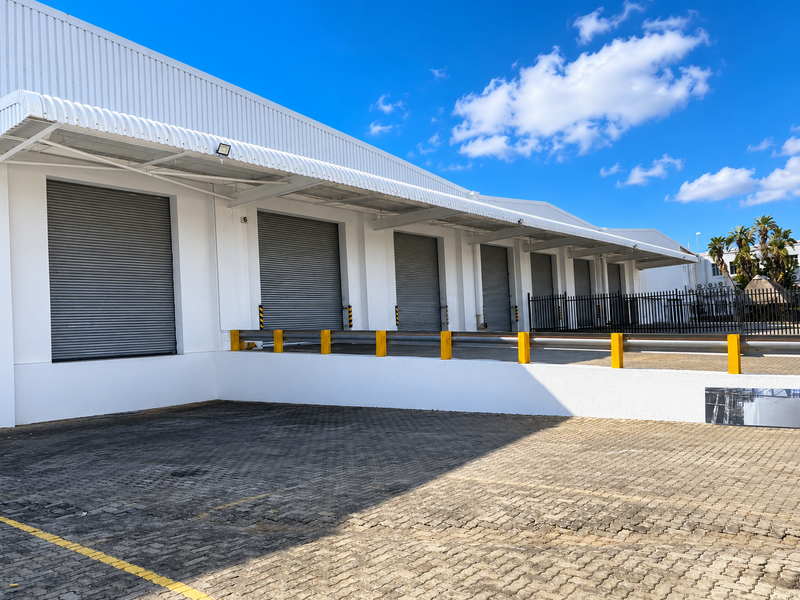Industrial warehouse exterior featuring multiple roller shutter doors, a raised loading bay with safety barriers, and a paved yard area.