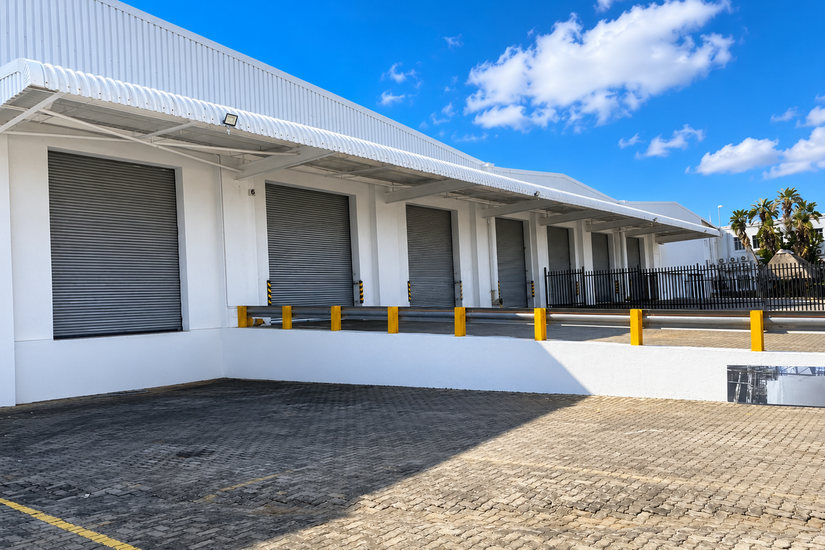 Industrial warehouse exterior featuring multiple roller shutter doors, a raised loading bay with safety barriers, and a paved yard area.