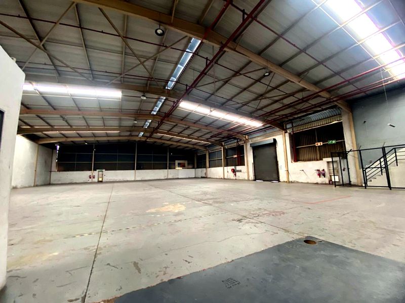 Spacious industrial warehouse featuring high steel truss ceilings, natural light from skylights, concrete flooring, and a large roller shutter door for easy access.
