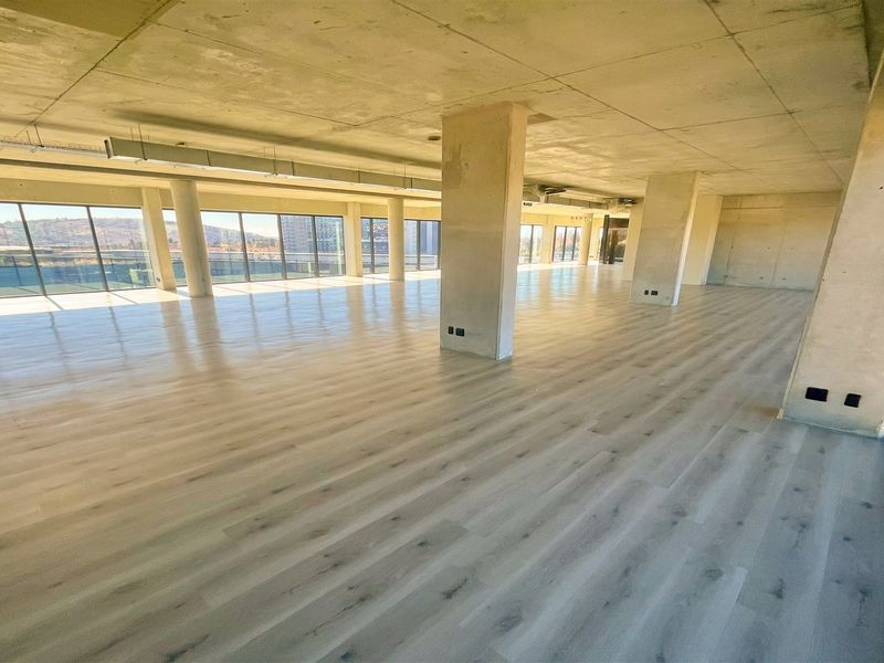 A spacious, open-plan office area featuring modern laminate flooring, exposed concrete ceilings, and expansive floor-to-ceiling windows providing ample natural light.